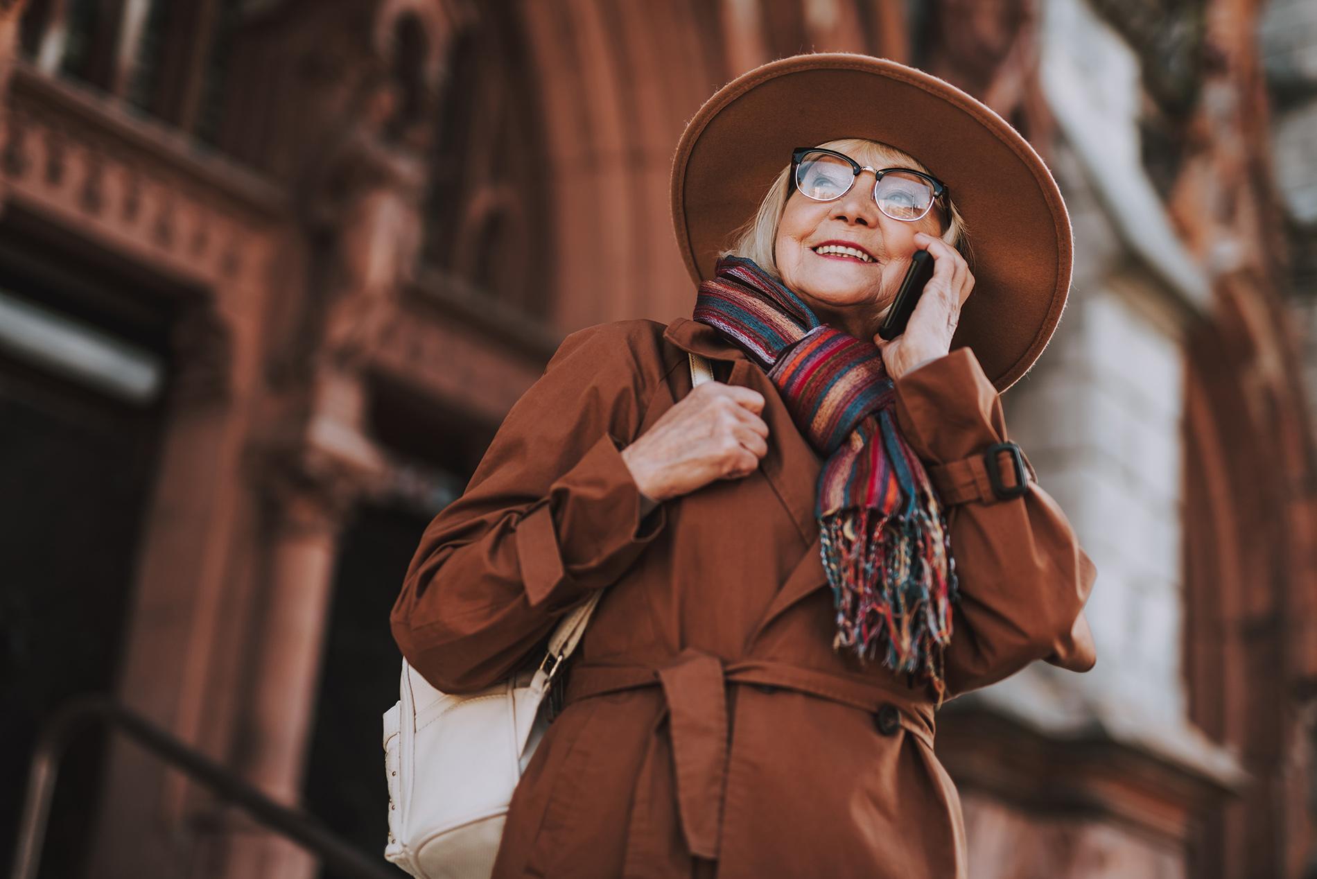 Lächelnde ältere Frau mit Brille im braunen Mantel telefoniert mit einem Handy.