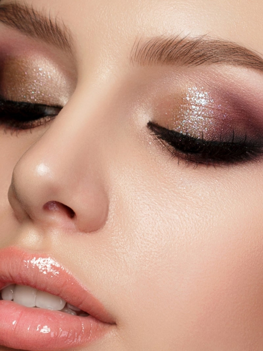 Ein Nahaufnahme-Porträt, das ein ausdrucksstarkes Augen-Make-up mit glitzerndem Lidschatten in Braun- und Violetttönen sowie glänzende, volle Lippen zeigt. Dieses Bild hebt die Kunst des Make-ups hervor und wie es das Gesicht zum Strahlen bringen kann. Es ist perfekt, um die Bedeutung von Details im Styling zu betonen und Inspiration für besondere Anlässe oder einen glamourösen Look zu bieten.