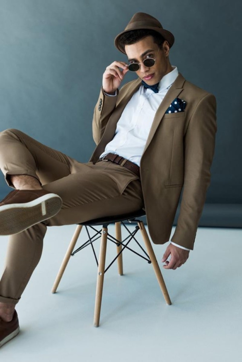 Ein stilvoller Mann in einem braunen Anzug, weißem Hemd, Fliege und runder Sonnenbrille, lässig auf einem Stuhl sitzend. Sein Look ist eine moderne Interpretation des 'Dapper'-Stils, der Eleganz mit einer entspannten Attitüde verbindet. Dieses Bild zeigt, wie man klassische Herrenmode mit individuellen Akzenten wie einer Fliege und markanten Accessoires aufwerten kann, um einen einzigartigen und selbstbewussten Auftritt zu erzielen.