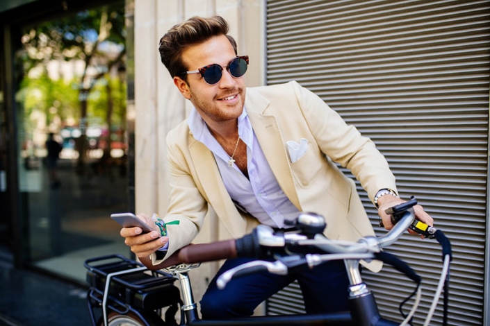Junger Mann mit modernem Outfit und Sonnenbrille auf dem Fahrrad – individuelle Fashion- und Stilberatung in Hannover. Ein stilvoller Mann lehnt lässig an seinem Fahrrad, gekleidet in einen beigefarbenen Blazer, ein hellblaues Hemd und eine dunkle Hose, ergänzt durch eine Sonnenbrille. Dieses Bild verkörpert den modernen Casual-Chic-Look für Herren, der sowohl entspannt als auch gepflegt wirkt. Es ist perfekt für den urbanen Alltag und zeigt, wie man auch auf dem Weg zur Arbeit oder in der Freizeit stilvoll unterwegs sein kann, ohne auf Komfort zu verzichten.