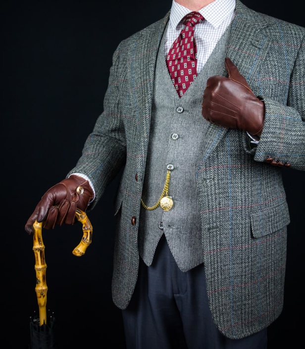 Ein Detailbild eines Mannes in einem klassischen Tweed-Sakko, einer passenden Weste und einem karierten Hemd mit roter Krawatte. Braune Lederhandschuhe und eine goldene Taschenuhr runden den Look ab, während er einen Spazierstock hält. Dieses Bild steht für zeitlose Eleganz und die Liebe zum Detail in der Herrenmode. Es zeigt, wie traditionelle Stoffe und Accessoires einen distinguierten und raffinierten Stil kreieren können